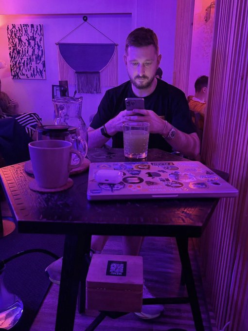 A man sits at a cafe table illuminated by purple lighting, looking down at his phone, with a sticker-covered laptop, coffee cup, and water glass in front of him, and a small box with a QR code on the floor.