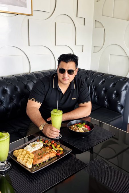 A man wearing sunglasses and a black "BOSS" polo shirt sits at a restaurant table with a green smoothie, a bowl of food, and a plate featuring salmon with grilled pineapple and rice, while the accompanying "GOOD MORNING Everyone - Touch this post to get rich in this September" caption appears unrelated to the meal, instead being a typical engagement-bait social media post.