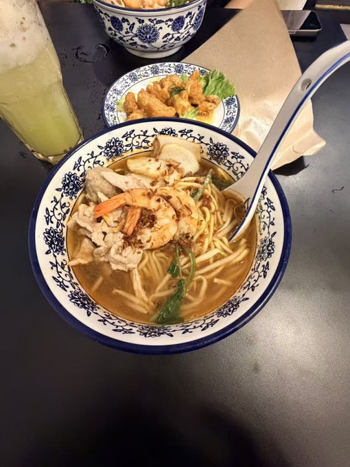A bowl of noodle soup with shrimp and vegetables, accompanied by a side of fried items, a green drink, and blue and white patterned dishware.