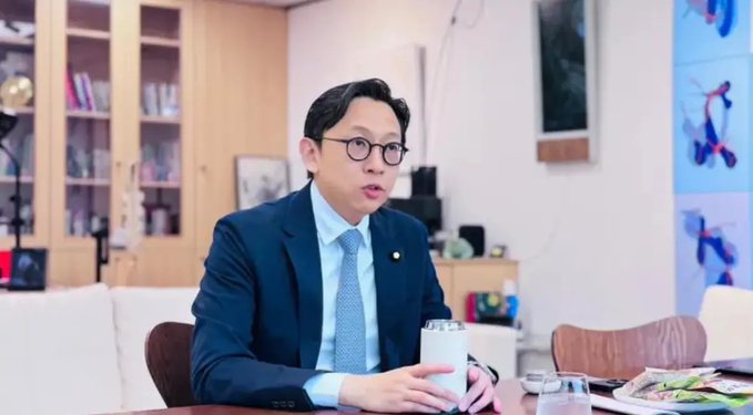 Dr. Ko Ju-chun, a Taiwanese legislator wearing glasses, a navy suit, and light blue tie, sits at a desk holding a white cup in what appears to be an office or study with bookshelves and artwork in the background, as he discusses Taiwan's potential strategic Bitcoin reserve.