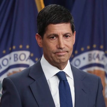 A man in a dark blue suit and tie stands before a backdrop featuring the seal of the Board of Governors, appearing to be at a Federal Reserve press briefing.