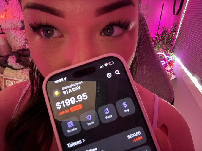 A close-up selfie of a woman holding a smartphone displaying a crypto wallet app showing a balance of $199.95 for the token '@alteregicotcpanic $1 A DAY', with pink ambient lighting in the background.