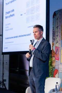 A man in a suit speaks into a microphone while presenting in front of a screen showing a slide about 'Cheaper code. More software.'
