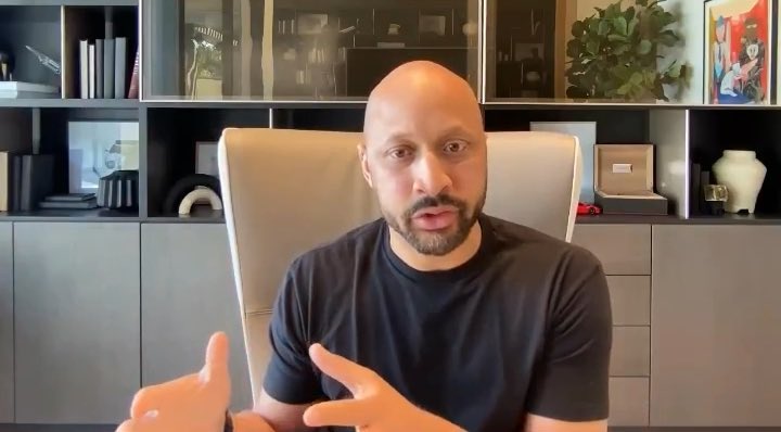 A bald man with a beard wearing a black t-shirt, sitting in an office chair and gesturing while speaking directly to the camera.