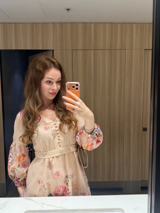 A woman with wavy brown hair takes a mirror selfie wearing a floral dress with puffy sleeves, holding a smartphone in what appears to be a restroom with wooden doors.