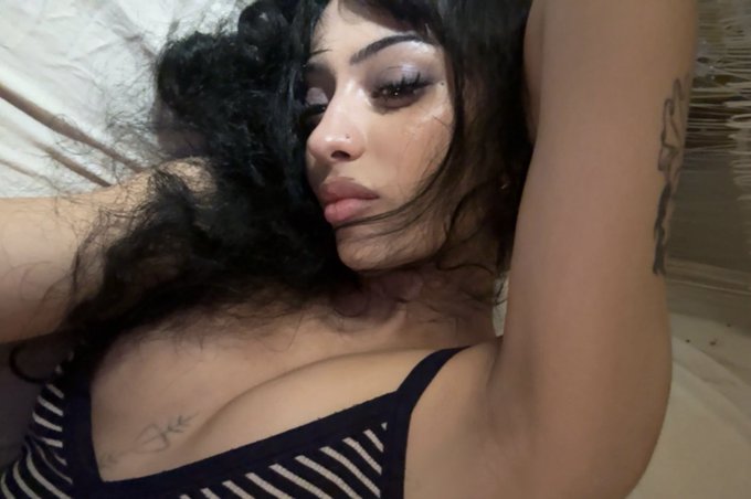 A young woman with long, curly black hair and dramatic makeup poses with her arm raised above her head, wearing a black and white striped top. Tattoos are visible on her chest and upper arm as she gazes at the camera against a softly lit background.