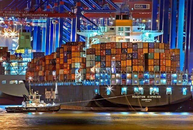 The image shows a Hapag-Lloyd container ship named "Houston Express" (registered in Hamburg) being assisted by a tugboat at night at a container terminal (Terminal Burchardkai, with gantry cranes marked "HHLA"), with its hull stacked high with colorful shipping containers—a vivid contrast to the dry bulk loading processes described in the post, which rely on grab buckets and conveyor systems rather than container cranes.