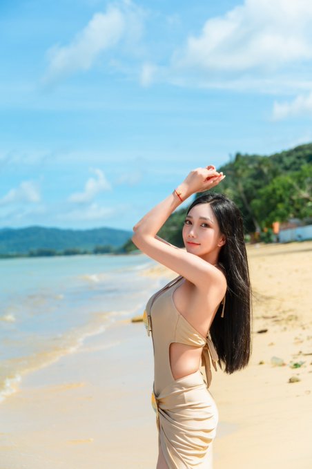 A young woman with long dark hair poses on a sandy beach in a tan/beige dress with side cutouts, with turquoise water, green mountains, and a blue cloudy sky in the background.