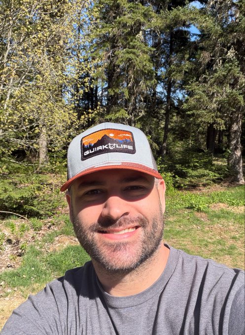 The image features a man wearing an orange and black baseball cap with the word "quirkiesnft" displayed on it, standing outdoors. The man has just taken a photo of himself and seems to be engaging with viewers by replying to comments or messages from them. The image promotes a product, specifically an item related to sports, with the name of the game being QuirkiesnFT. There is also text that references streamers on Twitch TV who might want to check out this particular item for themselves, possibly indicating its usefulness among fellow gamers.
