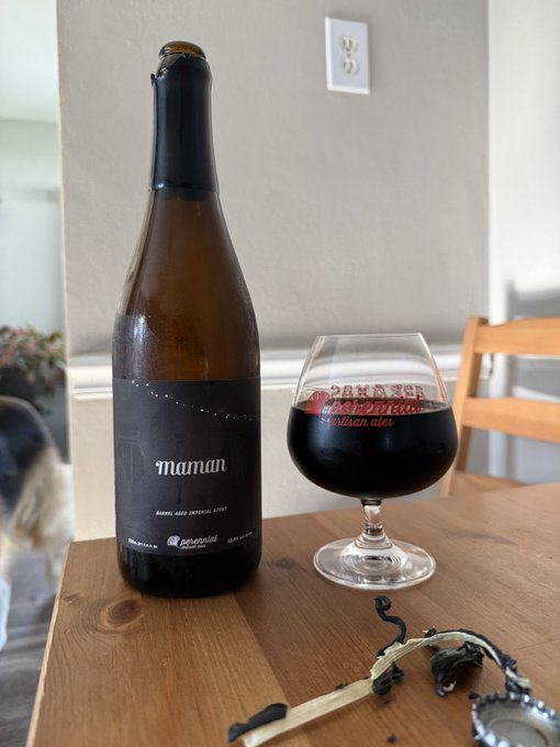 A bottle of 'maman' barley-aged imperial stout beer from Pipeworks Brewing next to a filled snifter glass on a wooden table, with a dog visible in the background.