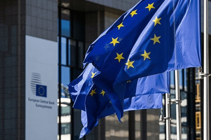 European Union flags fly outside the European Central Bank headquarters, which displays signage reading "European Central Bank Eurosystem," as MiCA crypto regulations take full effect across EU member states.