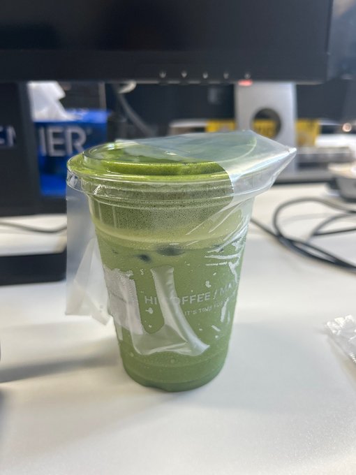 A plastic cup of iced matcha latte with a sealed clear plastic cover sits on a desk in front of a computer monitor, with "HI COFFEE / M" visible on the cup.