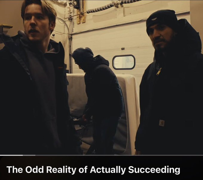 Photo of two men in winter clothing in a garage/workshop setting with the caption 'The Odd Reality of Actually Succeeding'.