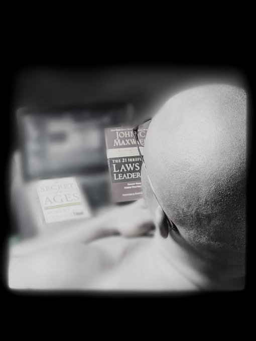 A close-up black and white photo of a bald person reading books titled 'THE 21 IRREVERSIBLE LAWS OF LEADERSHIP' by John C. Maxwell and 'SECRET AGES'.