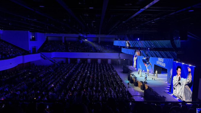A wide shot of a concert arena with performers on stage and large screens displaying "DAOU OFFROAD" to a large seated audience.