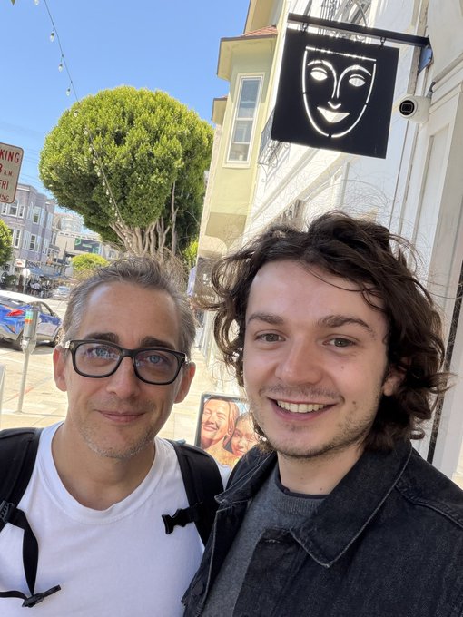 Two smiling men pose for a selfie on a sunny street in San Francisco, with a black sign featuring a stylized white face design hanging behind them. The casual, candid shot captures a moment between what appears to be the post's author and Mike Merrill, the "publicly traded person" referenced in the caption, visually tying the creator token pioneer to the story being shared.