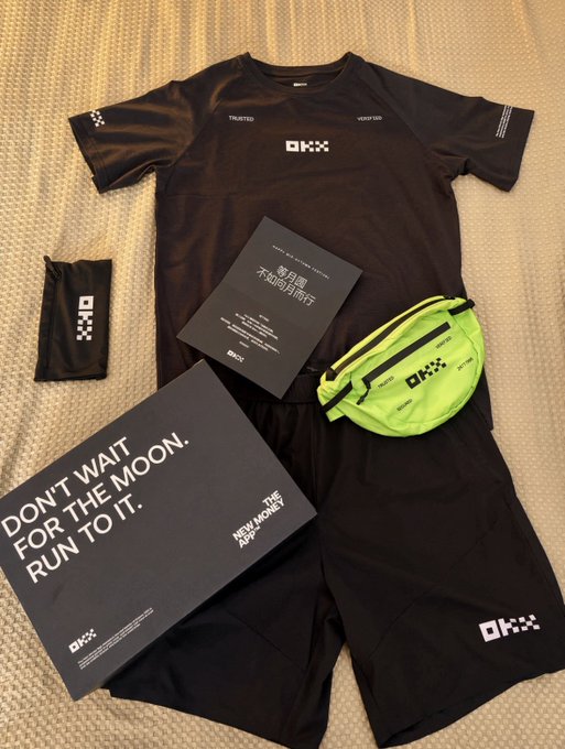 A flat-lay of black OKX-branded running gear including a 'TRUSTED / VERIFIED' shirt and shorts, a green neon belt bag, an instruction booklet, and a box reading 'DON'T WAIT FOR THE MOON. RUN TO IT.' with the 'NEW MOON' logo.