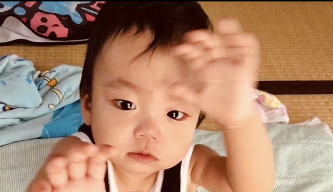 A close-up portrait of a young baby with dark hair reaching their hand toward the camera.