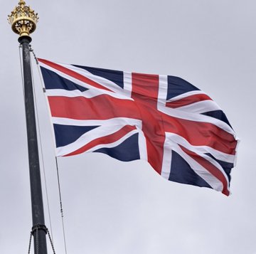 The British Union Jack flag waves against an overcast sky, symbolizing the UK's forward momentum as it officially launches applications for crypto licenses under its new regulatory framework—positioning itself ahead of the US, which has stalled on the CLARITY Act. The image visually reinforces the post's message about the UK taking the lead in crypto regulation while other nations lag behind.