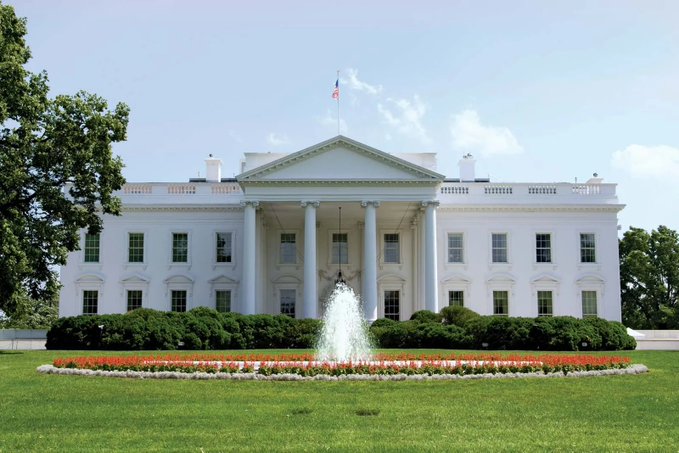 The White House stands prominently with its iconic north portico columns and a flag flying above, set against a backdrop of manicured gardens and a central fountain. This historic backdrop underscores the significance of today's unprecedented crypto summit, where President Trump is set to convene with top regulators like SEC Chair Paul Atkins and CFTC Chair Mike Selig alongside industry leaders from Coinbase, Ripple, Gemini, and Polymarket.