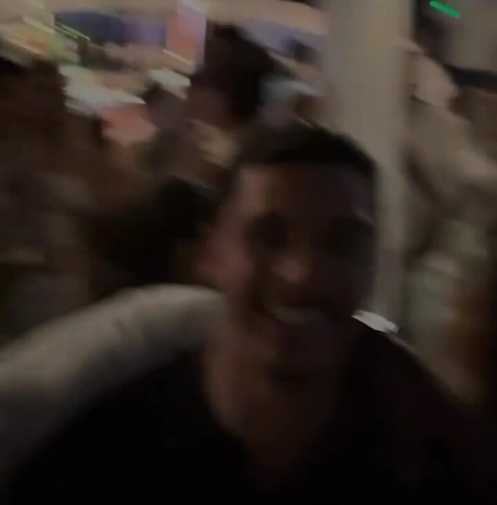 The image shows a man with black hair wearing a black shirt sitting down at a table. The background appears blurry, suggesting that he might be indoors or moving quickly. There are other people present in the scene as well, but they are not the main focus of the image.