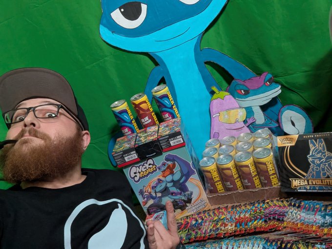 A person with a reddish beard wearing glasses and a black cap, sitting in front of a green backdrop with a large SNEK character (blue frog-like mascot) and Pokemon characters, surrounded by SNEK energy drink cans and product boxes.