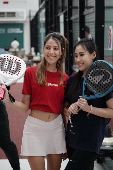 The image features two young women standing next to each other holding tennis rackets. They are both smiling for a photo, showcasing their enthusiasm for the sport. The background of the image shows a group of people gathered around, indicating that they might be at an event or gathering related to tennis or sports in general.