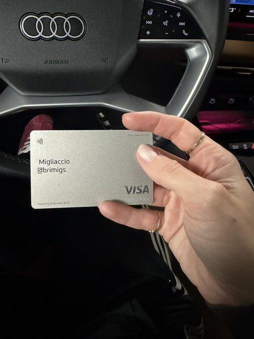 A hand holding a gray Visa debit card with the name 'Migliaccio @brimigs' is shown inside a car, with the Audi logo visible on the steering wheel.
