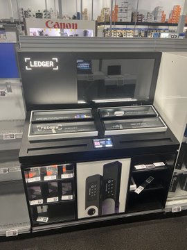 The image shows a display of wallets at a store, with two different types available for purchase - a Ledger wallet and a Tangem wallet. The wallets are placed on a black shelf or countertop, making them easily visible to customers. The store also offers additional products such as cell phones, which can be found in the background. A sign is displayed above the display, advertising the availability of these items at the store for sale.
