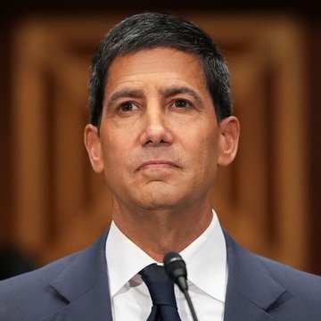 A serious-looking man in a dark suit and tie stands before a wooden backdrop with a microphone visible, matching the accompanying context about Fed Chair Kevin Warsh preparing to deliver his Jackson Hole speech on Friday at 10:00 AM ET, where markets await signals on inflation and interest rate policy.