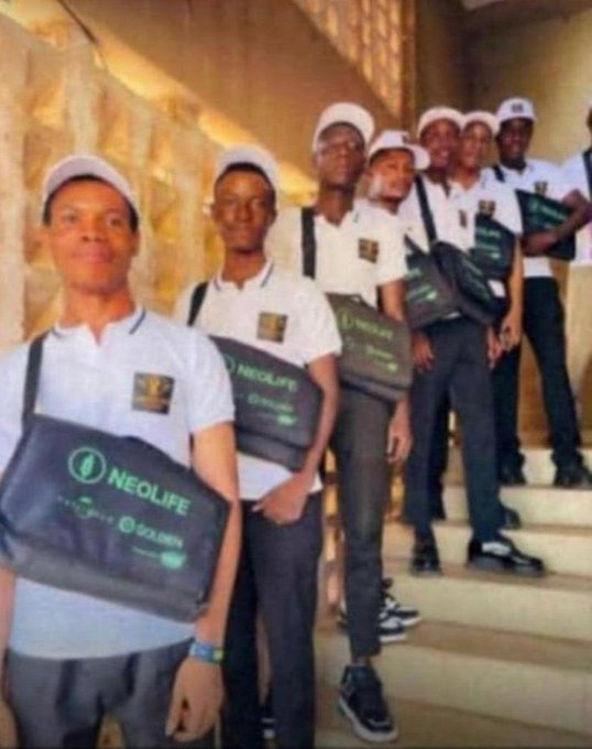 A group of young men in matching white polo shirts and caps standing on stairs, each carrying black bags branded with the 'NEOLIFE GOLDEN' logo.
