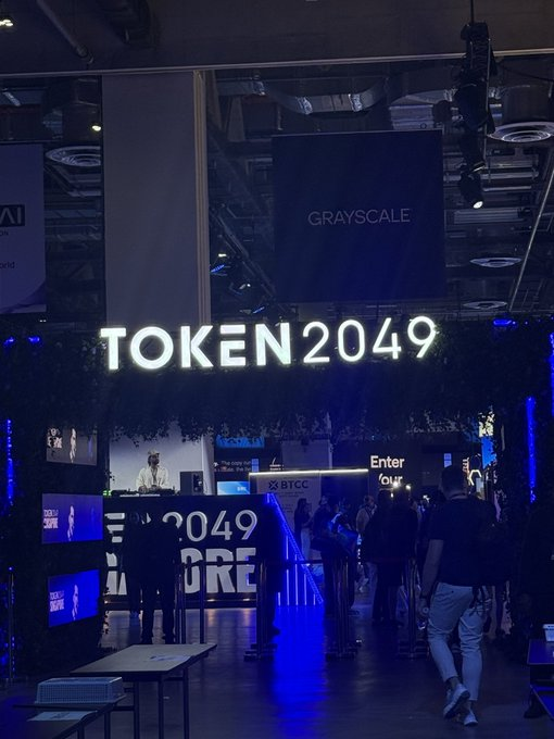 An entrance to the TOKEN2049 crypto conference, with a large illuminated 'TOKEN2049' sign flanked by Greyscale and BTCC logos, where attendees are walking into the blue-lit venue.