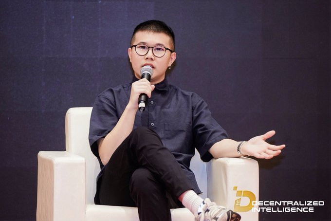A young man with glasses and a buzz cut sits in a white chair on stage, holding a microphone and speaking; the logo "Decentralized Intelligence" appears in the corner.