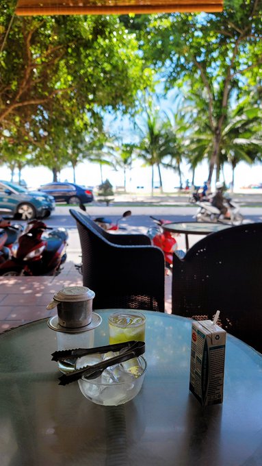 A table at an outdoor beachside café holds a small pot, a glass of iced drink, and a carton, with motorbikes, palm trees, and the ocean visible in the background.