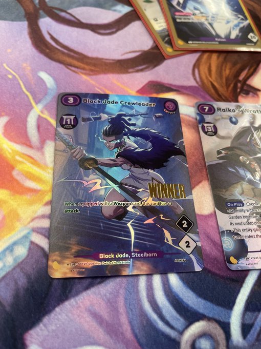 Trading cards on a playmat, prominently featuring a 'Black Jade Crewleader' card with 'WINNER' text and 'Black Jade, Steelborn' subtitle, alongside a 'Raiko' card.