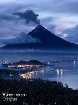 A scenic nighttime photograph of a volcano with glowing red lava erupting, overlooking a coastal town with illuminated bridges and water reflections, with Chinese text crediting '新华社' (Xinhua News) and photographer 黎寒冰.