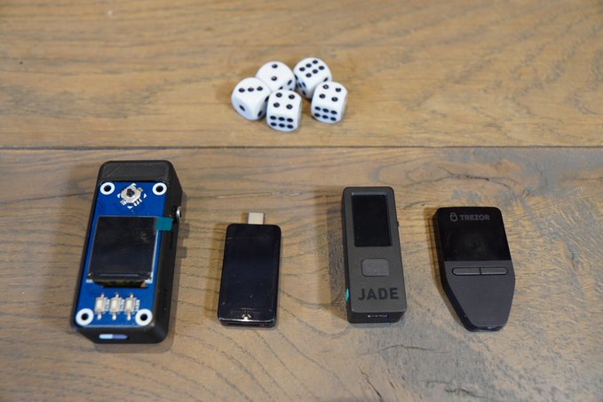 Four hardware cryptocurrency wallets displayed on a wooden table next to a cluster of white dice, including brands like Trezor and Jade.