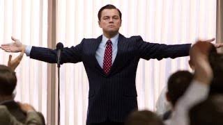 A man in a pinstripe suit and red tie stands with arms outstretched in front of an applauding audience, reminiscent of a Leonardo DiCaprio Wolf of Wall Street pose.