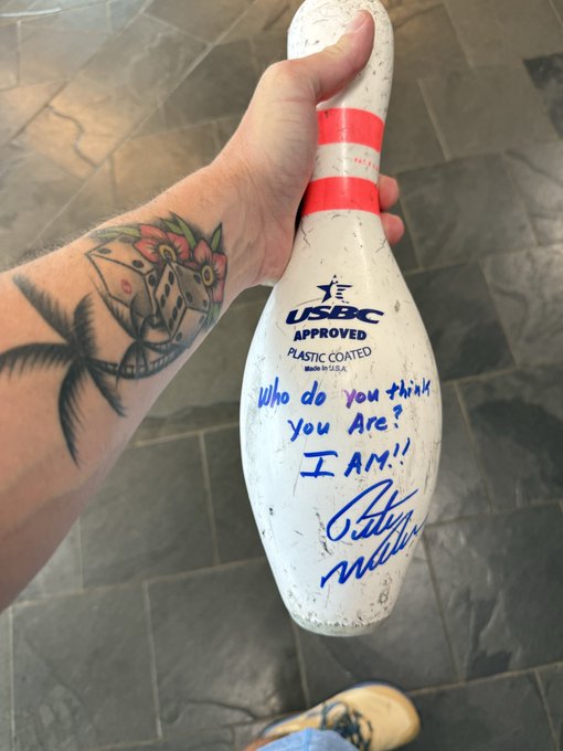 A tattooed hand holds a well-worn USBC-approved plastic-coated bowling pin autographed with the inscription "Who do you think you are? I AM!" — a personal "grail" piece commemorating a meaningful milestone.
