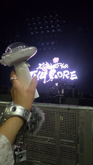 A concertgoer holds up a heart-shaped K-pop lightstick (resembling those from the group ILLIT) at a live music event, with a brightly lit stage screen reading "ILLIT " in the background.