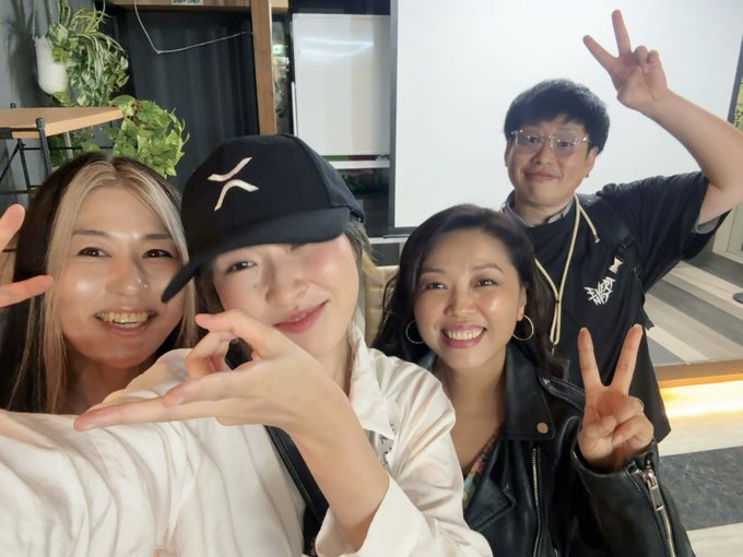 A selfie of four people posing indoors; a woman on the left and a woman on the right are making peace signs, while a woman in the center wearing a black cap makes a heart gesture with her hand, and a man in the back right holds up a peace sign.