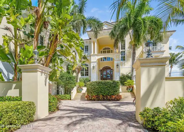 A large yellow Mediterranean-style luxury home with palm trees, a paved driveway entrance, and lush landscaping under a bright blue sky, marked with '26 Space Coast MLS' watermark.