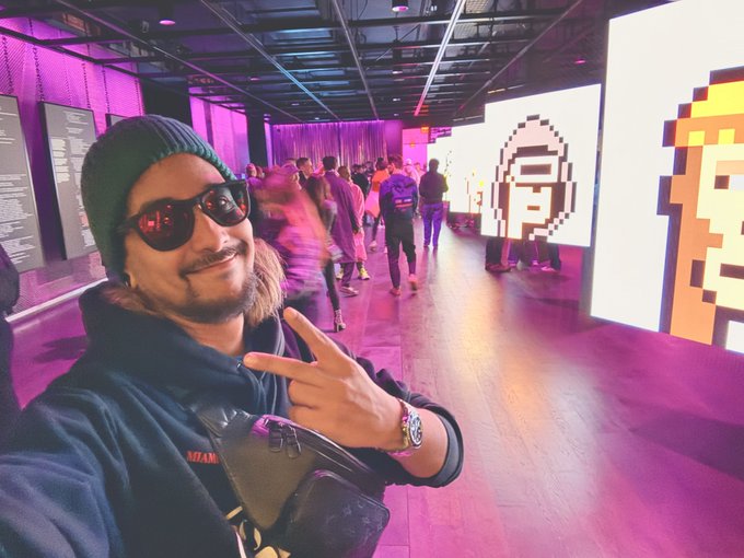A man wearing a green beanie and red-tinted sunglasses taking a selfie while making a peace sign at what appears to be a colorful crypto or NFT event with pixel art displays.