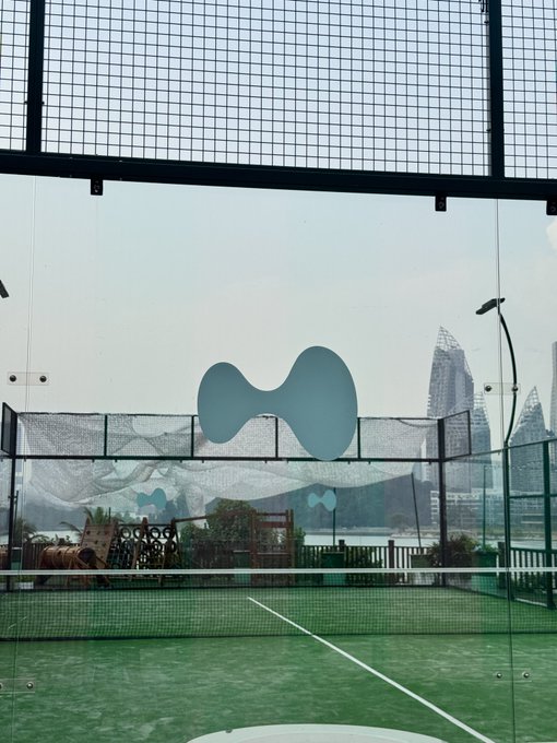 A paddle tennis court with a blue logo on the glass enclosure, overlooking a body of water and city skyline.