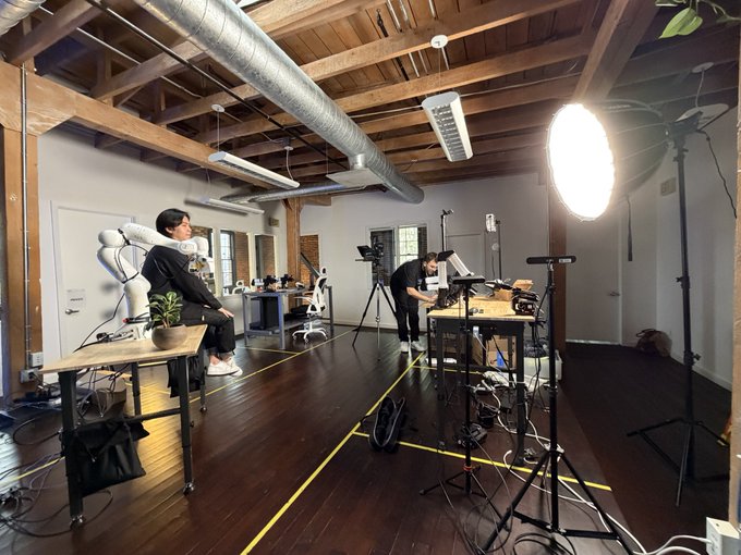 Behind-the-scenes view of a video production setup in a loft space, showing a person sitting on a chair in front of a green screen while crew members adjust cameras and lighting equipment.