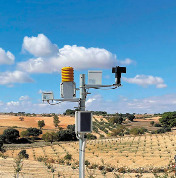 An agricultural weather station with various sensors and a yellow component stands in a rural landscape under a blue sky with scattered clouds.