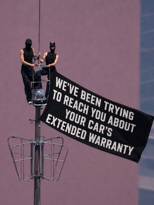 Two masked figures on top of a street pole unfurling a banner that reads 'WE'VE BEEN TRYING TO REACH YOU ABOUT YOUR CAR'S EXTENDED WARRANTY'.