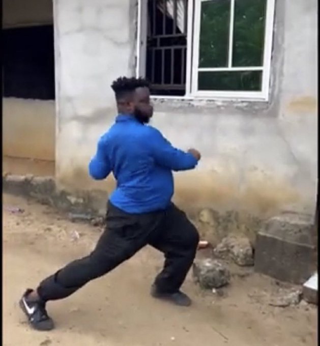 A man in a blue shirt and black pants is captured in a wide stance, appearing to dance or move energetically on a dirt ground next to a building.