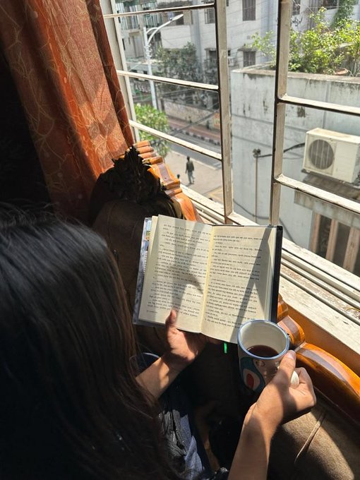 A person reads a book while holding a cup of tea by a window overlooking a quiet street, enjoying a peaceful morning ritual. The cozy, reflective moment complements the post's theme of starting the day with mindful engagement and earning rewards through staking in the River ecosystem.