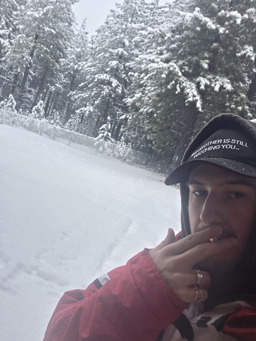 A selfie of a man in a red jacket smoking a cigarette outdoors in a snowy landscape, wearing a black cap that reads 'THE KILLER IS STILL WATCHING YOU.'